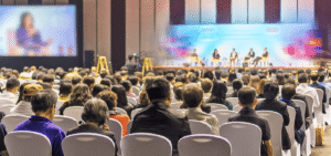 Audience listening to a panel discussion on stage at a conference, with multiple speakers and a large screen on the left.