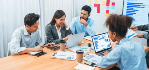 Team of four professionals collaborating around a table with laptops and charts in a bright meeting room.