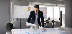 Businessman in a navy blazer reviews documents spread across a conference table with charts and graphs in an open office setting.